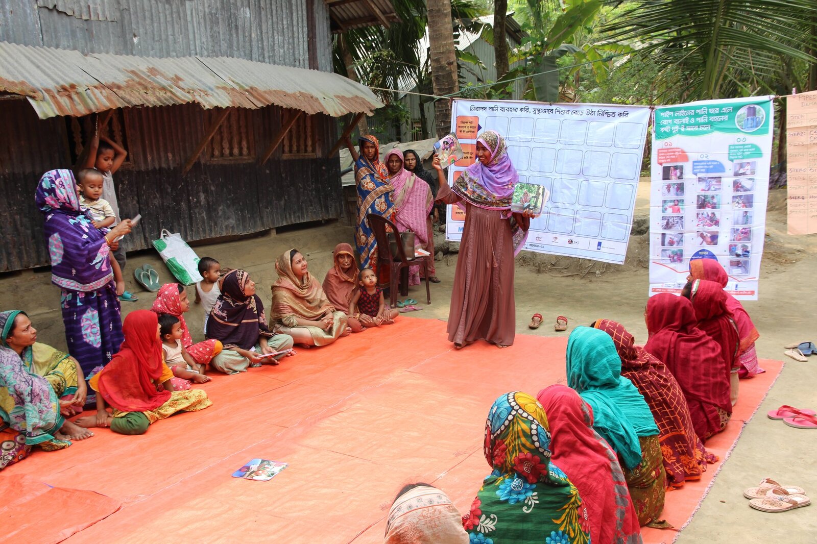 Women and children seated at an outdoor courtyard session on safe water and hygiene practice
